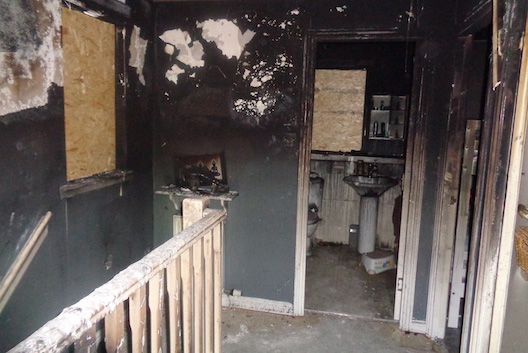 Fire-damaged upper hallway and bathroom of house, with blackened walls and appliances and boarded-up windows.