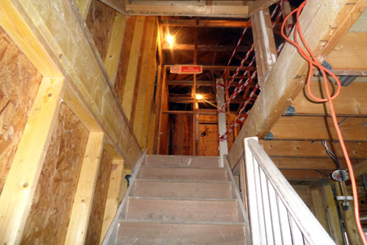View from stairway showing scaffolding on upper floor with wooden walls, exposed flooring, cabling, red and white safety tape and no smoking sign.