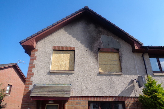 External view of fire-damaged house with boarded-up windows and damaged wall and roof on upper floor. House Fire Damage Reinstatement Airth case study.