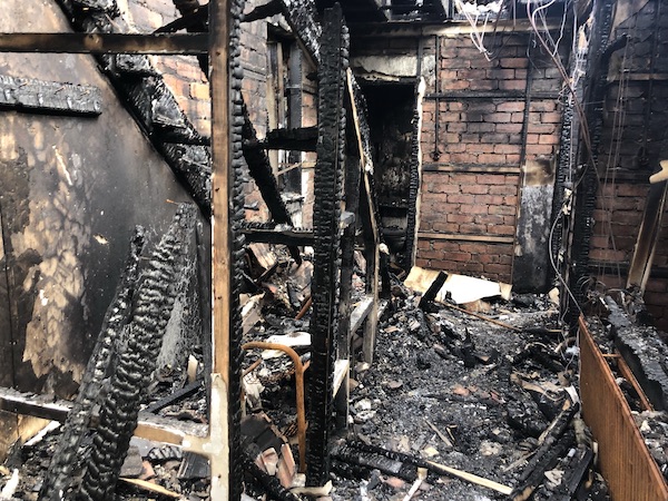 Internal view of fire-damaged property with blackened brickwork, exposed wooden beams and tarnished metalwork.