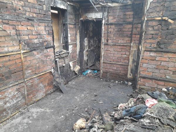 Internal view of property with bare brick walls, exposed wooden beams and doorway, dangling metal and debris on the ground.