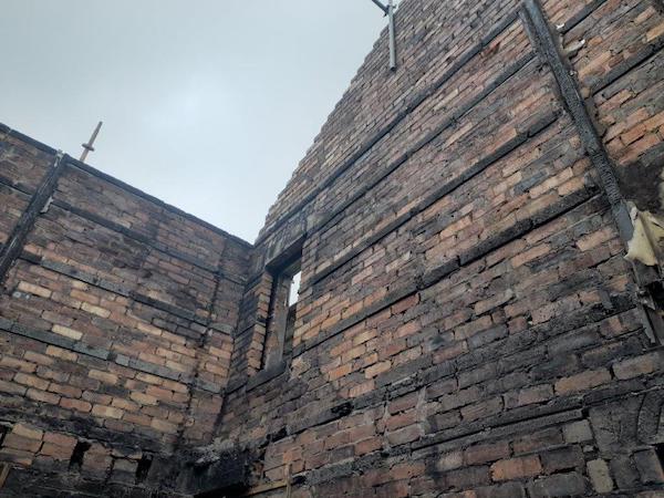 Exposed brickwork with blackened wooden beams on walls and empty window frame with vertical scaffold legs at top of the walls.