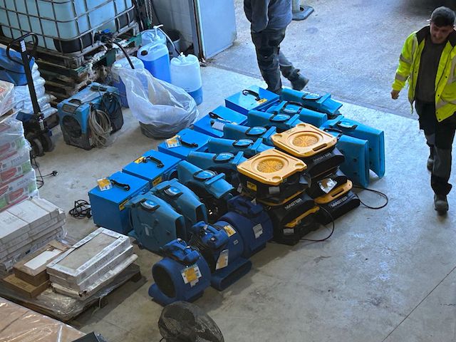 A range of industrial drying equipment and other items on a warehouse floor. A workman in a yellow high-vis jacket walks past. blog surge and flood