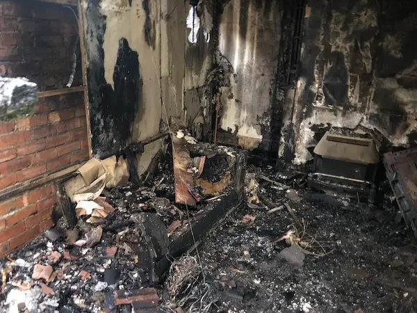 Internal view of fire-damaged room with exposed brickwork and beams, blackened walls, damaged fireplace, couch and TV.